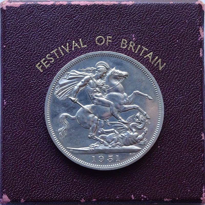 Silver Crown coin of 1951, produced for the Festival of Britain, showing a helmeted and plumed, naked Saint George on horseback, facing right, cloak flying behind him, attacking a dragon with his sword. The date, 1951, is in the exergue, and the coin lies on top of its box, which is dark red, and the text, Festival of Britain, is visible.